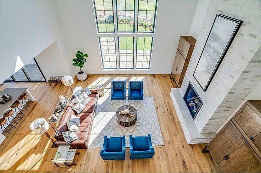 这是迷人的农舍风格客厅的俯视图，高高的天花板配上高高的玻璃窗。客厅的家具包括一张棕色沙发和两对蓝色扶手椅，围绕着壁炉对面的一张木制咖啡桌。