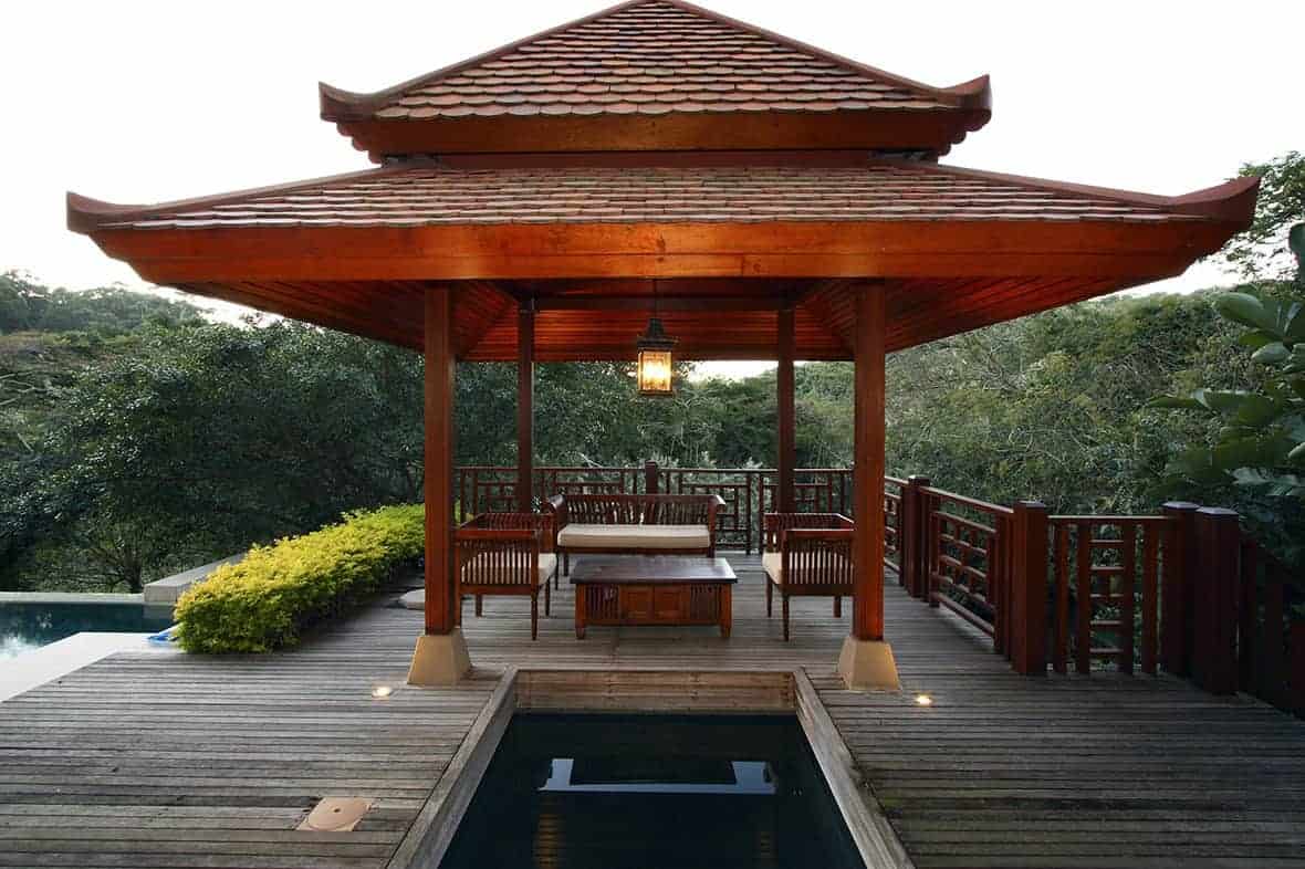 这个亚洲风格的木制凉亭的屋顶具有明显的中国设计，红色的色调与柱子一致，两侧的木栏杆以及下面的木沙发由一个巨大的灯笼状吊灯照亮。