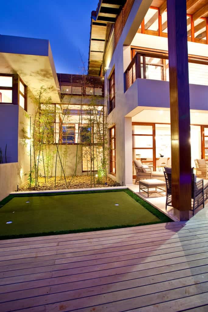 这座明亮的亚洲风格住宅有一个室外露台，露台上铺着木地板，配有配套的木椅和凳子，凳子上有白色靠垫。旁边是一个小高尔夫球区，有三个洞，背景是高大的竹林和黄色的聚光灯。