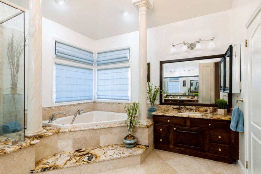 地中海风格的主浴室，铺着瓷砖地板，玻璃窗上覆盖着浅蓝色的卷帘。它包括一个深色的木质梳妆台和角落里镶有大理石柱子的浴缸。