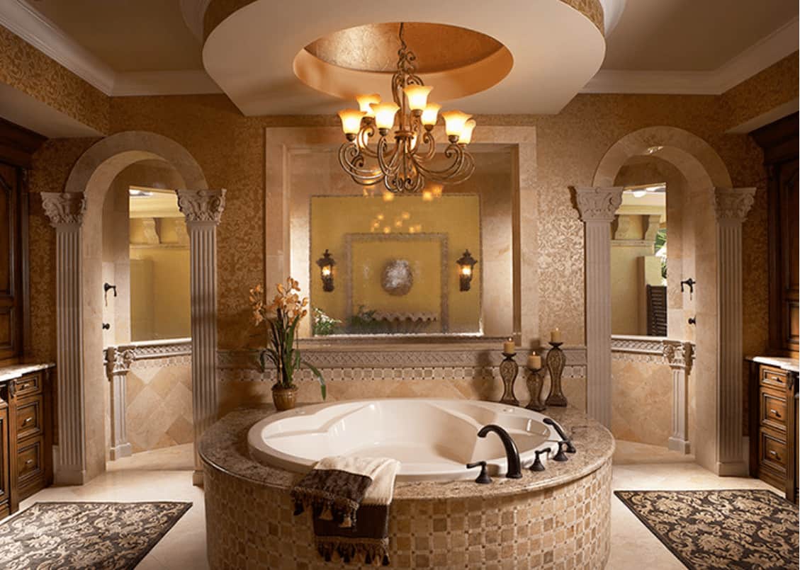 优雅的主浴室，圆形浴缸被悬挂在托盘天花板上的华丽吊灯照亮。里面有漂亮的地毯和通往步入式淋浴间的拱形门。