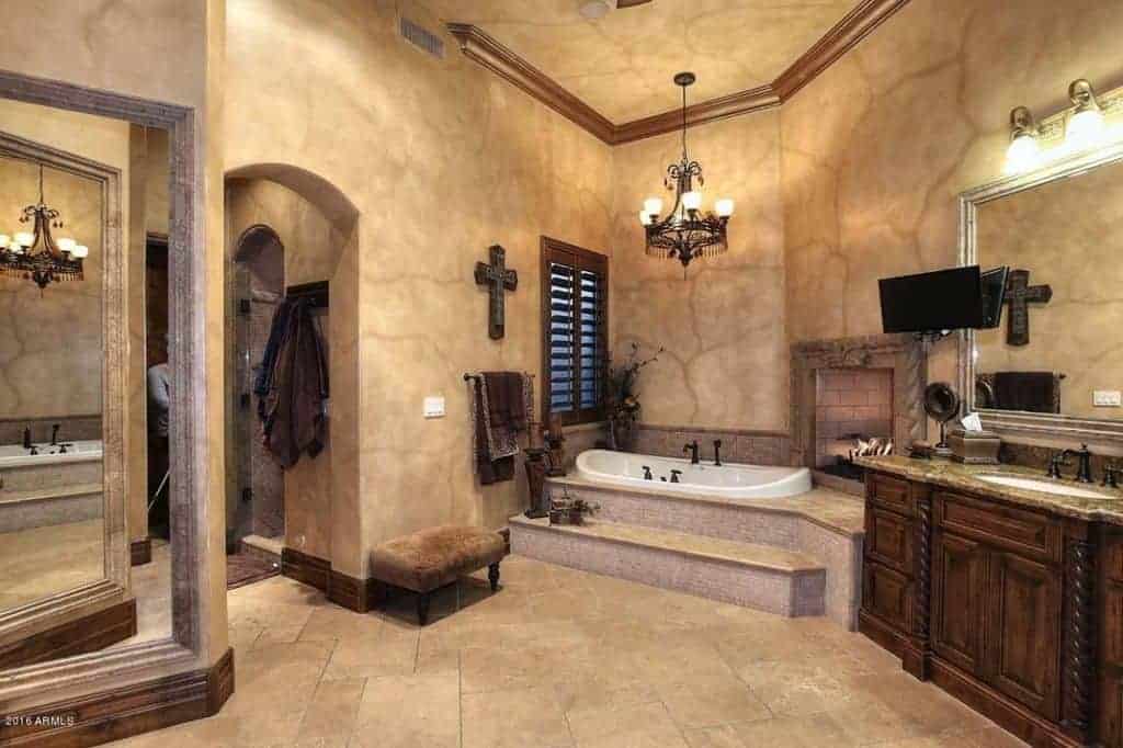 地中海风格的主浴室，带有洗手台和固定在浴缸上方的砖砌壁炉。它由一盏老式的枝形吊灯照亮，它悬挂在常规的天花板上，内衬是木制的
