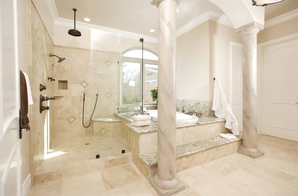 地中海风格的主浴室，带有步入式淋浴间和浴缸，浴缸里有大柱子。它铺着瓷砖地板，有一扇拱形窗户，可以让自然光照进来。