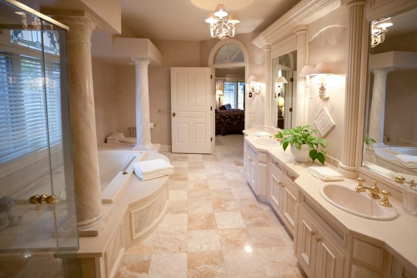 一扇白色木门通往这间主浴室，里面有一个落地式浴缸和双洗手池梳妆台，配有黄铜固定装置和五金设备。它由一盏枝形吊灯和安装在米黄色墙壁上的配套烛台照明。