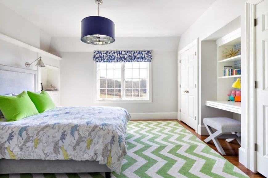 这个孩子卧室的彩色床单和枕头与绿白色图案的地毯很好地搭配在一起，地毯主要覆盖了硬木地板。窗户的蓝色图案阴影和白色天花板上悬挂的装饰性吊灯的蓝色兜帽增强了这些效果。