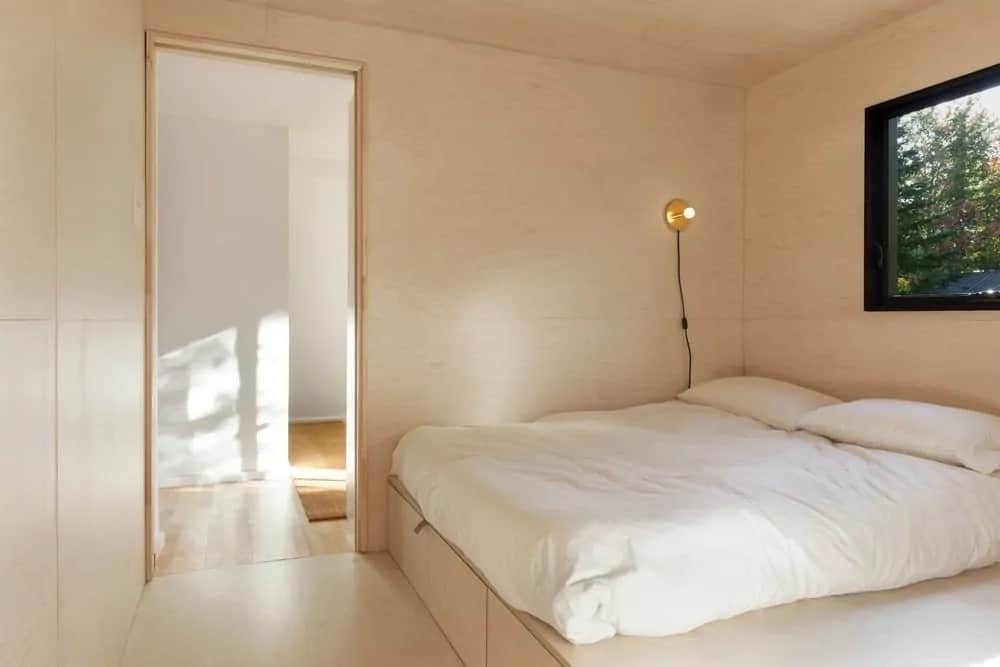 这间简单的村舍风格卧室的地板、墙壁、天花板甚至平台床的框架都搭配了浅色的木质调。它有一个白色的靠垫，补充了灯光色调和从上面窗户进来的自然光线。