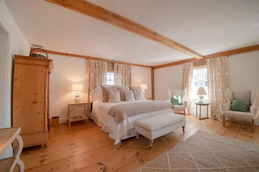 这间小屋风格的主卧室的天花板和墙壁上裸露的木梁与硬木地板和传统床旁的大木柜完美匹配，传统床旁有软垫床头板和脚处的软垫长椅。
