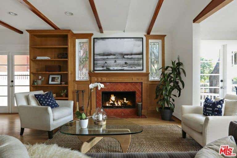 小屋风格的客厅拥有一个壁炉，上面有黑白相间的艺术品，还有一个玻璃顶咖啡桌，旁边是白色软垫椅子，深蓝色的枕头。