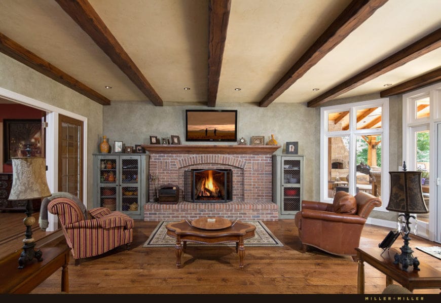 天花板上裸露的深色大木梁与硬木地板和漂亮的木制咖啡桌很相配。它位于两张不同的扶手椅之间，面对着巨大的红砖壁炉，电视安装在壁炉上方的墙上。