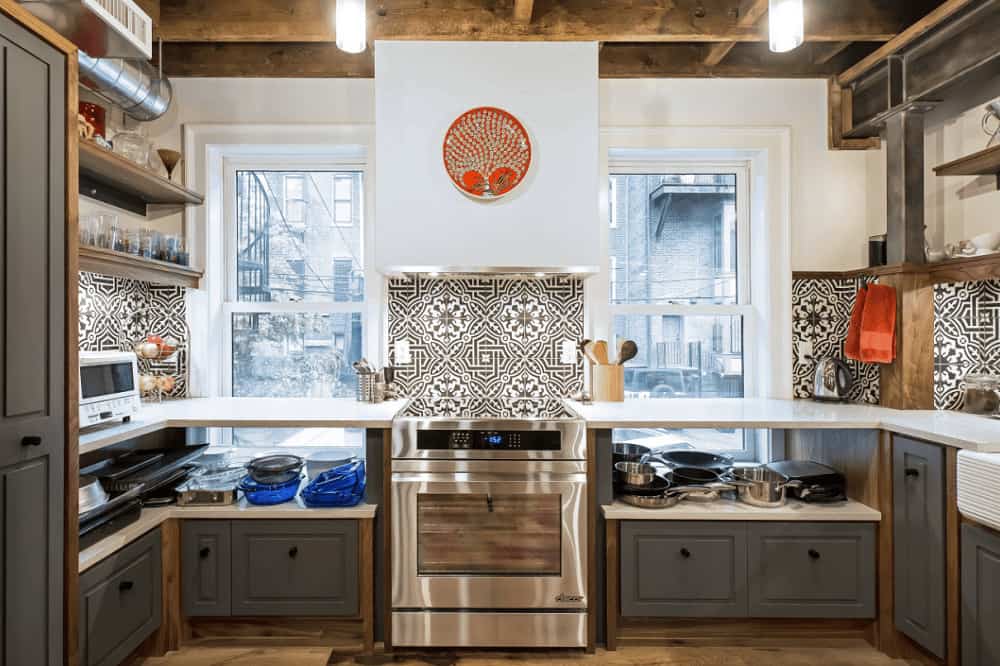 一对玻璃吊坠悬挂在木梁天花板上，照亮了这个厨房，展示了灰色橱柜和固定在白色石英台面上方的装饰性后挡板瓷砖。