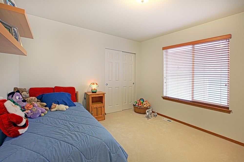 这是一个简单的儿童卧室，有一个宽敞的地毯地板空间供孩子玩耍。浅米色的墙壁与传统床脚上方的木制浮动架子和木制床头柜相辅相成，木制床头柜上装有一盏小灯。