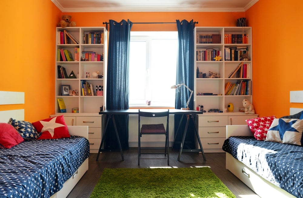 这个孩子的共享卧室里亮橙色的墙壁为两张白色的床奠定了欢快的基调，这两张床与靠窗的书桌两侧的白色书架相匹配。这带来了丰富的自然采光，使白色的天花板和绿色的地毯更加明亮。
