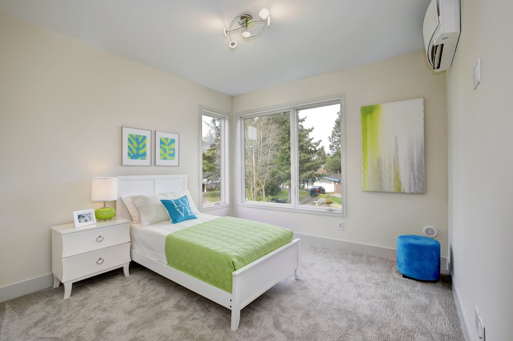 这间明亮宽敞的儿童卧室铺着灰色地毯，与传统的白色床和白色床头抽屉相映成趣。这与米色的墙壁很搭，墙壁上装饰着与床单相配的彩色抽象画。