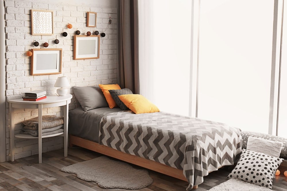 小床的灰色花纹床单与灰色砖墙和灰色色调的硬木地板很相配，白色床头柜附近铺着灰色地毯。宽阔的窗户使这些灰色元素变得明亮，并辅以微小的橙色细节。