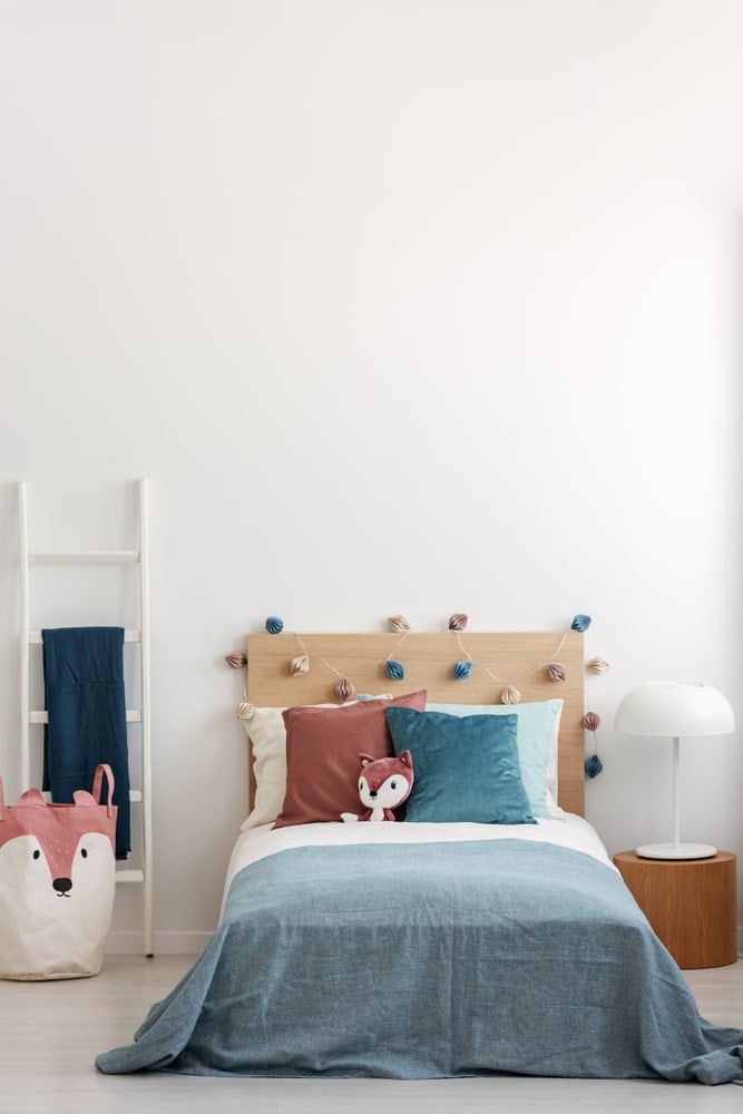 这是一个简单而小的孩子的卧室，有一个小平台床，有一个木制床头板，装饰着彩色的装饰品和枕头，以及一只毛绒狐狸。这与床旁的垃圾箱相匹配，既可以用来洗衣服，也可以用来放玩具。