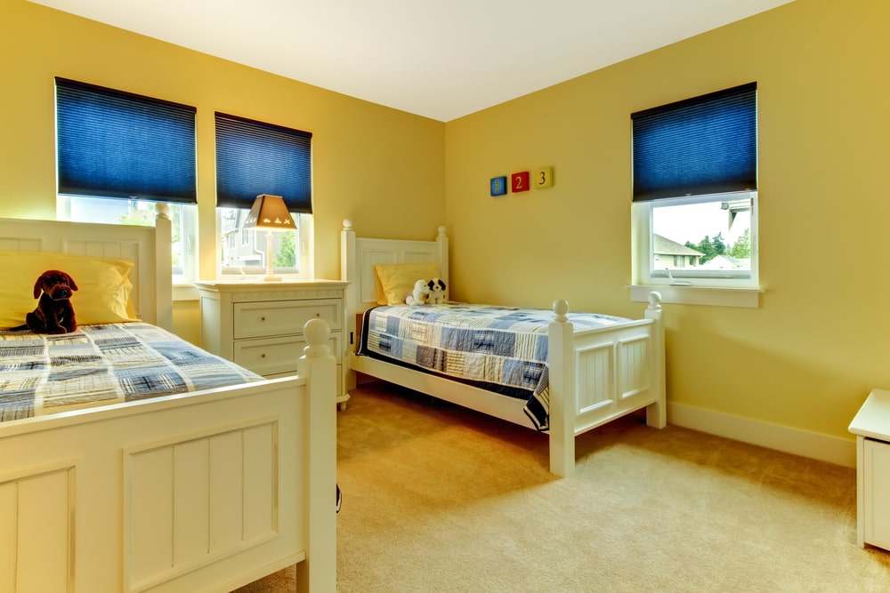 这是一间阳光明媚的孩子共享卧室，有两张黄色的木床，与芥末黄色的墙壁和米黄色的地毯地板相适应。这些床也与中间窗户下的梳妆台搭配，梳妆台上有一盏黄色的灯。