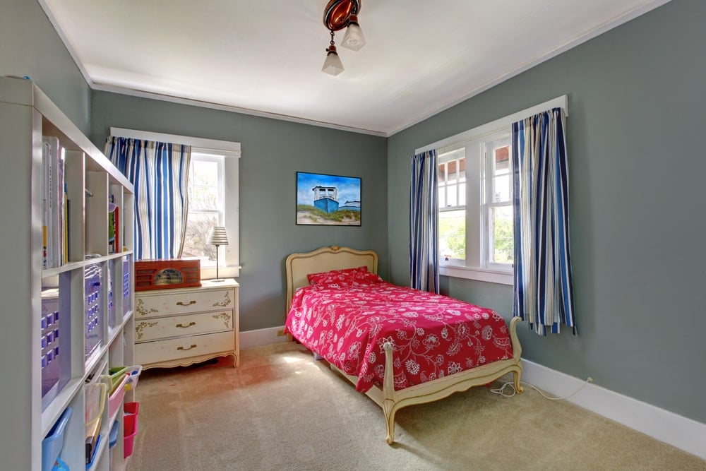 米色雪橇床的红色图案床单在这个孩子的卧室里很显眼，周围是灰色的墙壁，白色的天花板和米色的地毯地板。这张床与靠窗一侧的优雅梳妆台很相配，梳妆台上的蓝色条纹窗帘与窗户的颜色相同。