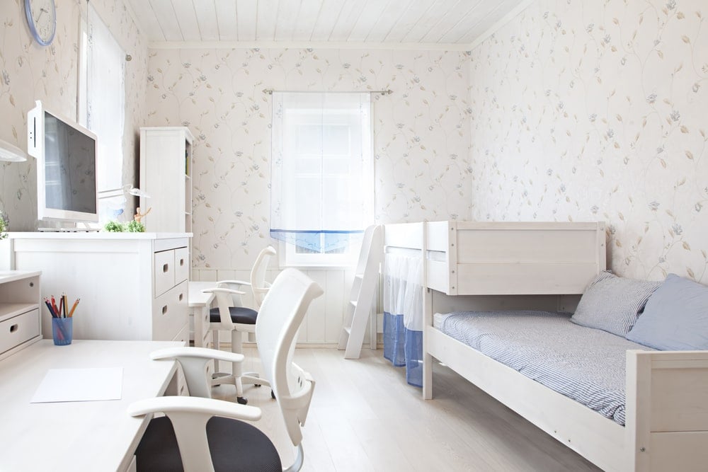 这个孩子卧室的亮度主要是由于充足的自然光从窗户射进来，照亮了白色图案的壁纸，有微小的细节。这些窗户也照亮了双层床、梳妆台和桌子的白色木质结构。