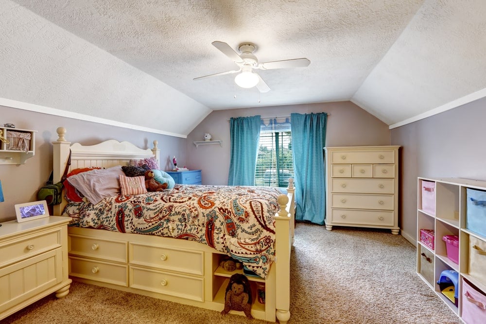 这间宽敞的儿童卧室有一个灰色的凹形天花板，与远处的窗户照亮的灰色墙壁很好地融合在一起。这种灰色的色调与黄色的木床、床边的抽屉以及窗边的大木制梳妆台相得益彰。