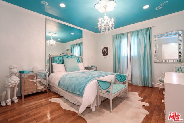 这间地中海风格的卧室的天花板是浅蓝色的色调，与床头板的蓝色靠垫和床脚的长凳相匹配。这也与窗户的蓝色窗帘相匹配，与浅灰色的墙壁和硬木地板形成对比。
