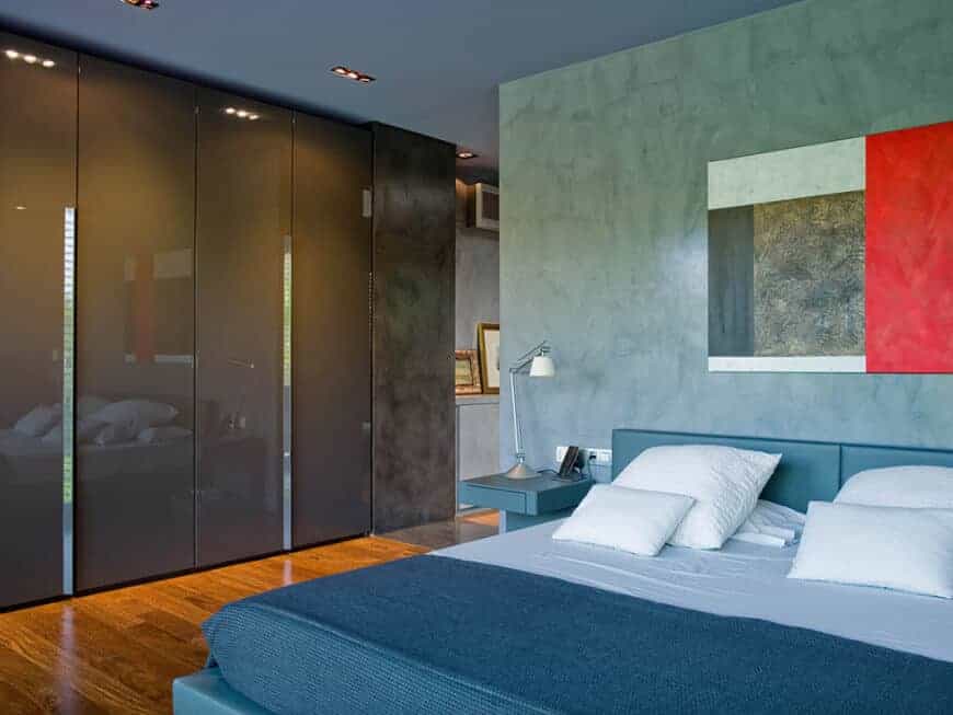 现代主卧室配有高光泽灰色衣柜和一张蓝色皮革床，并配有内置床头柜。混凝土墙上装饰着一件光滑的艺术品。