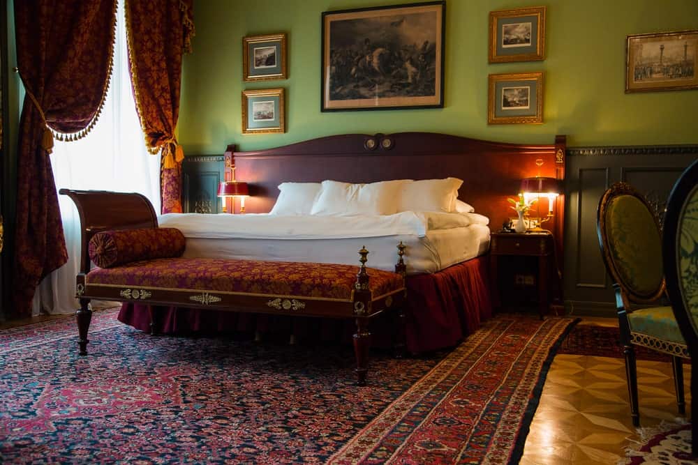 经典的主卧室，有一张带裙的床和优雅的躺椅，与红色图案的窗帘相辅相成。它包括一个花卉区域地毯和挂在绿色墙上的画廊框架。