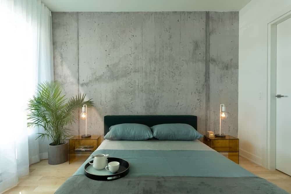 在这间工业风格的主卧室里，一盆棕榈树创造了一种清新的氛围，卧室里有一张靠混凝土强调墙放置的平台床。它的两侧是半透明的床头柜和灯泡台灯。