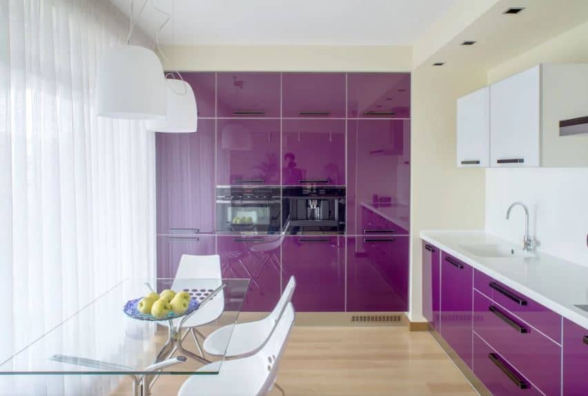 现代化的厨房也是餐厅的一部分，餐厅里有一张玻璃桌面的桌子和现代的白色椅子，这些椅子被大窗帘的窗户照亮。这些也点亮了嵌入厨房墙壁的光滑的深紫色橱柜。