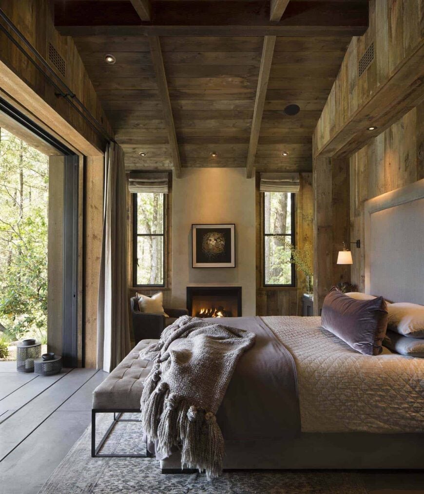 这个放松的乡村风格的主卧室最大限度地利用了美丽的户外风景，它的墙在床脚，打开。与此相邻的是一个壁炉，两侧是床旁的窗户。它们的顶部都有一个高高的木制教堂天花板，上面有暴露的木梁。