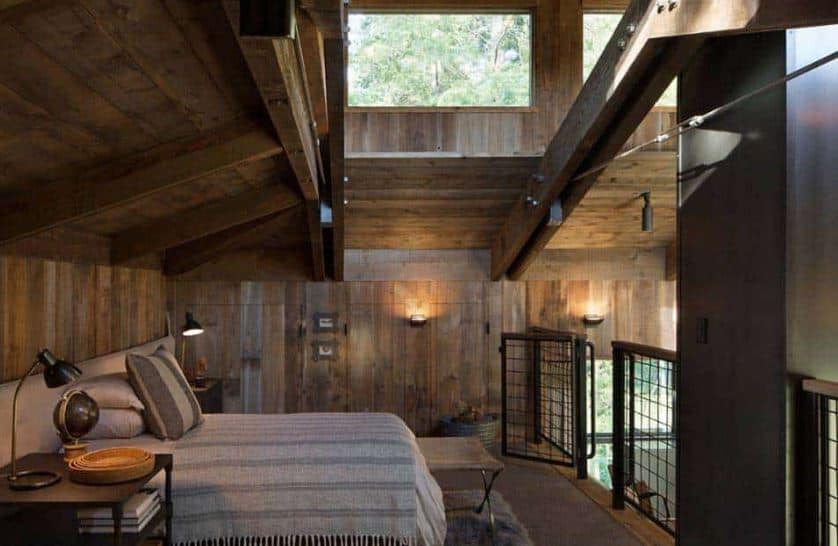 简单的灰色床、灰色床单和灰色床头板与木墙和露出木梁的天花板相得益彰。这些横梁和木制天花板被明亮的横窗照亮了。