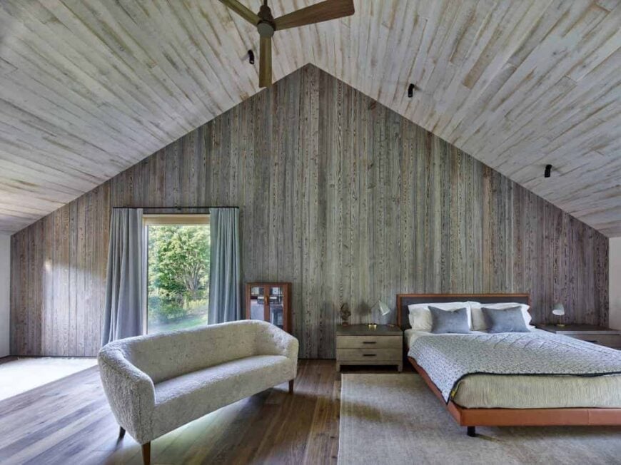 这间乡村风格的大卧室有一座大教堂式的木质天花板，与木墙和硬木地板融为一体。这包围了床和软垫沙发，与区域地毯相匹配，融入了迷人的木制元素。