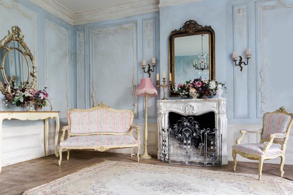 优雅的客厅装饰着华丽的镜子，挂在白色的控制台桌上，壁炉围在一个华丽的三面板屏幕上。它有一个破旧别致的区域地毯和迷人的座椅，由一盏粉红色的落地灯和老式的烛台照亮。