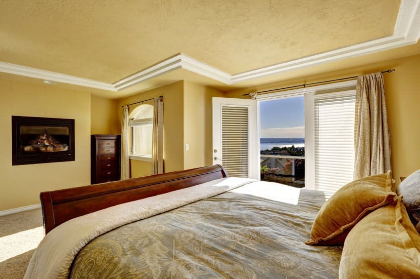 黄色的卧室带有拱形窗户和法式门，通往阳台，享有宁静的室外景色。房间里有一座壁挂式壁炉和一张木床，床上铺着柔和的蓝色花纹被褥。
