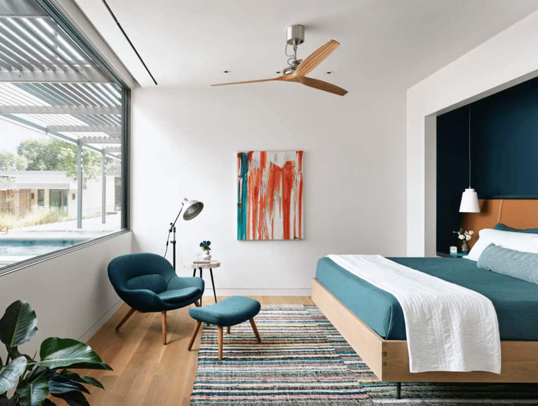 白色的主卧室，带有抽象艺术品和蓝绿色圆形靠背躺椅，与嵌入墙上的舒适床相映成辉。
