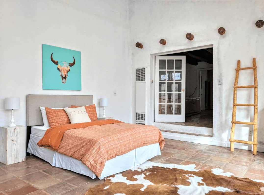 一个牛头艺术品为这个白色的卧室增添了一个不错的口音，灰色的裙子床和牛皮地毯铺在陶土地板上。