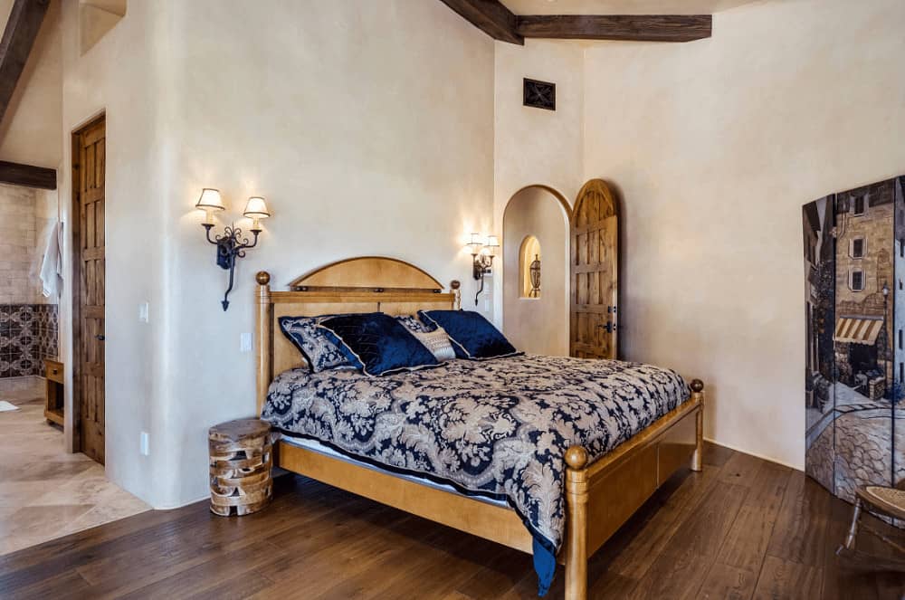 一扇拱形雕花木门通往这间舒适的卧室，里面有锻铁壁灯和一张木床，床上装饰着蓝色碎花床品。