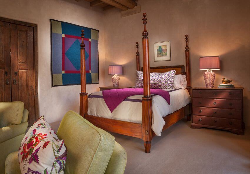 这间西南风格的卧室里有一张木制铅笔海报床，床单上有浅色，与挂毯的大胆色彩相匹配，挂毯挂在床边抽屉台灯旁边的白墙上，与床架的色调相匹配。