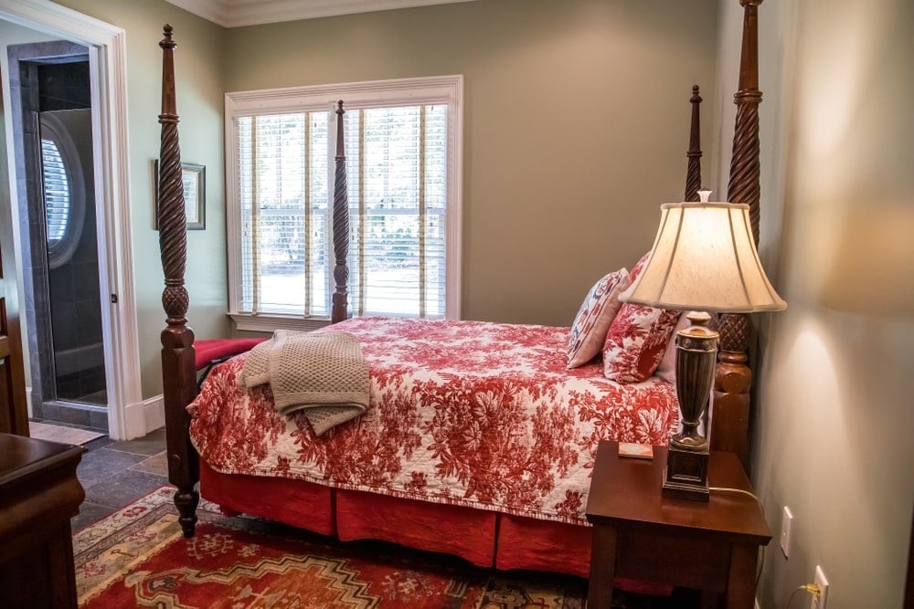 四柱床的红色花卉图案床单与下面的红色图案地毯相匹配。这主要包括西南风格卧室的泥土地板，辅以浅灰色的墙壁和带百叶窗的白色窗户。