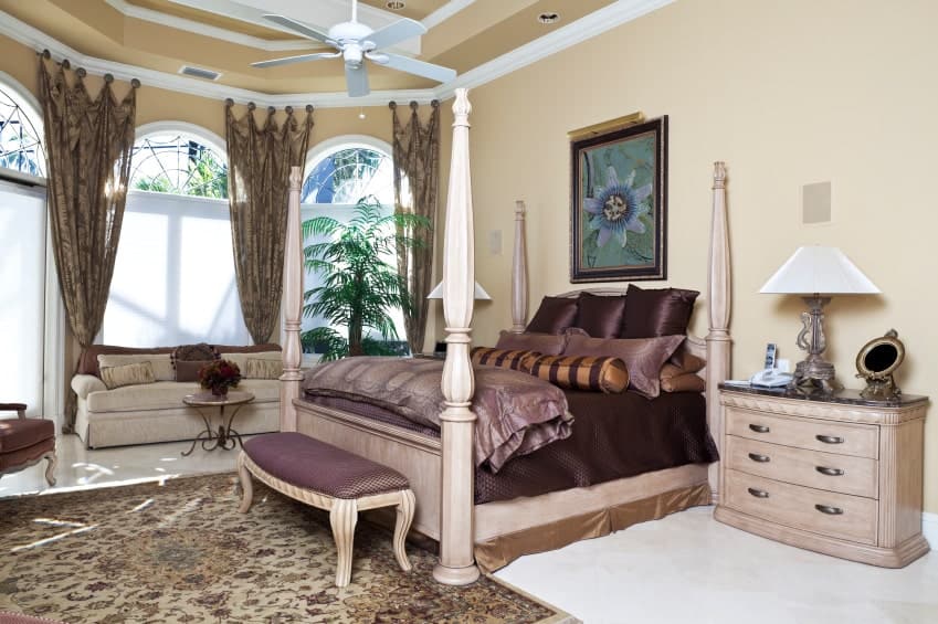 优雅的主卧室四海报展示了一个木制的床和一个米色的沙发的拱形窗户穿着热带窗帘。它包括花卉艺术墙和白色的风扇挂在天花板托盘。