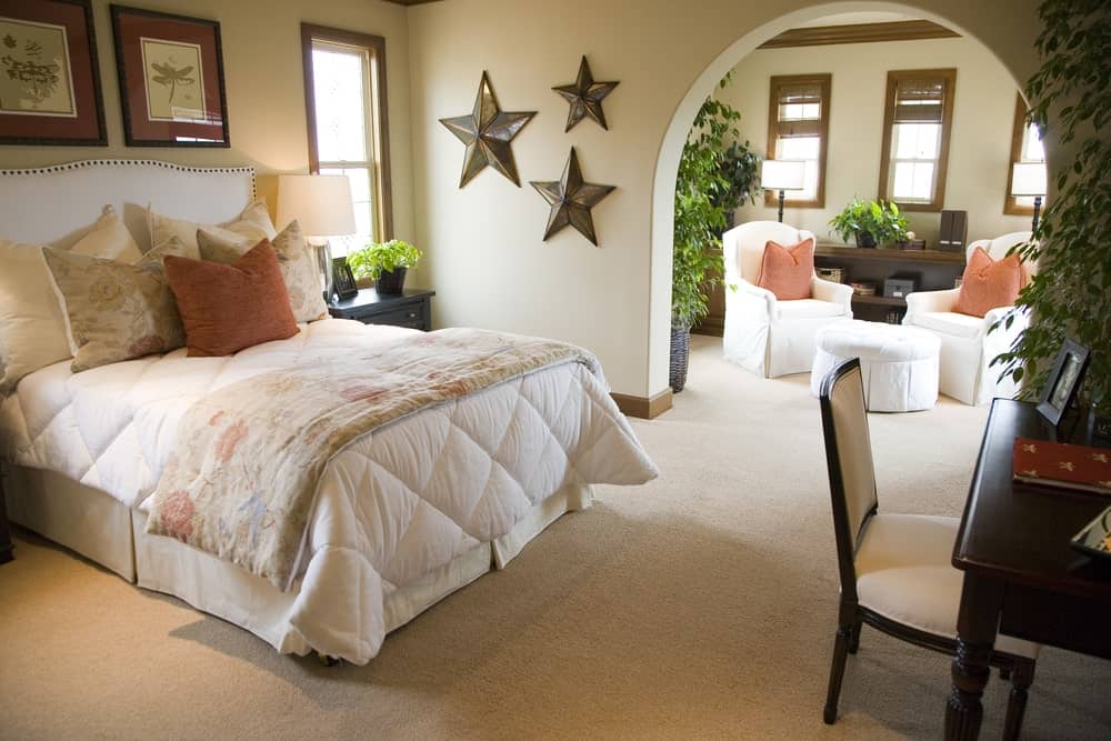 迷人的卧室地毯地板和一个开放的拱门导致座位区。明星艺术墙和白色的床上,面对黑暗的木桌子搭配米色柔软的椅子。