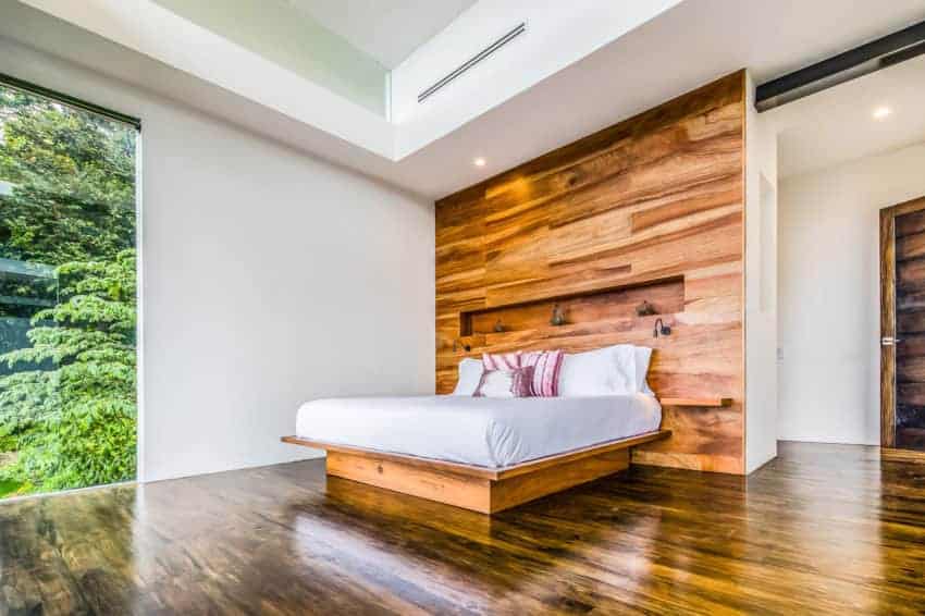 木制平台床,白色的垫子和枕头混合的大型木制墙壁作为大型床头板。这有一个红木色调,与白色的墙壁,白色的托盘天花板和玻璃窗。