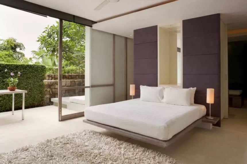 现代浮动平台床有一个白色靠垫,反衬着深灰色的床头板。白色的床是进一步改善自然灯光来自大型滑动玻璃门与热带树木和植物。