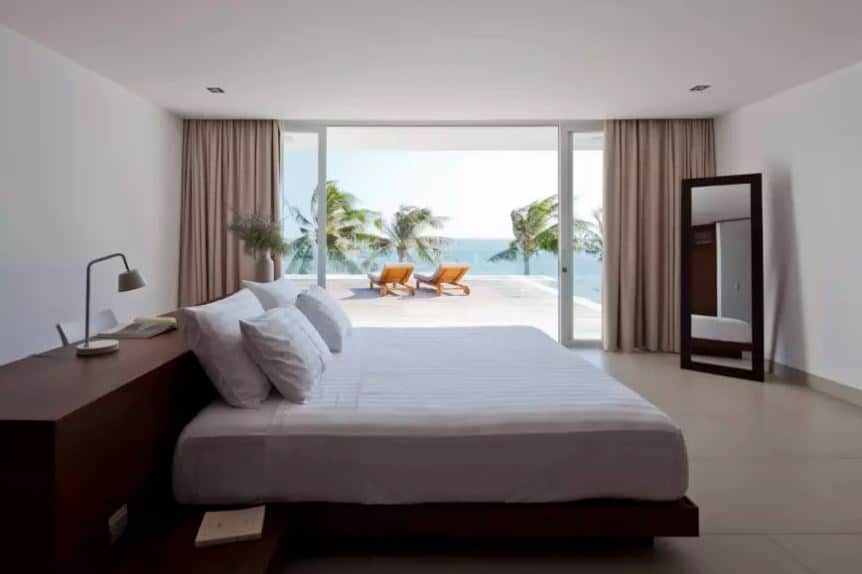 突出的热带风格卧室外面的景色壮丽,顶部的椰子树对海洋的观点。这是一个放松的视图对于那些正在享受简单的白色平台床上有一个很大的木质床头板。