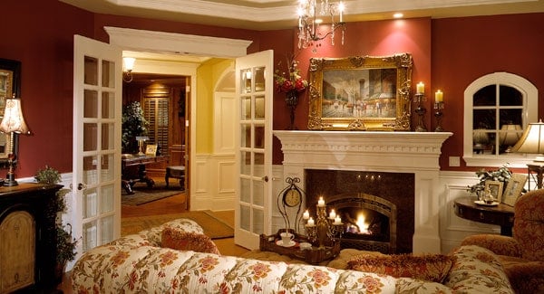 一扇白色的法式落地门通往客厅，客厅里有碎花座椅和一座温暖的壁炉，壁炉固定在红墙和碎花沙发对面的白色护壁板上。