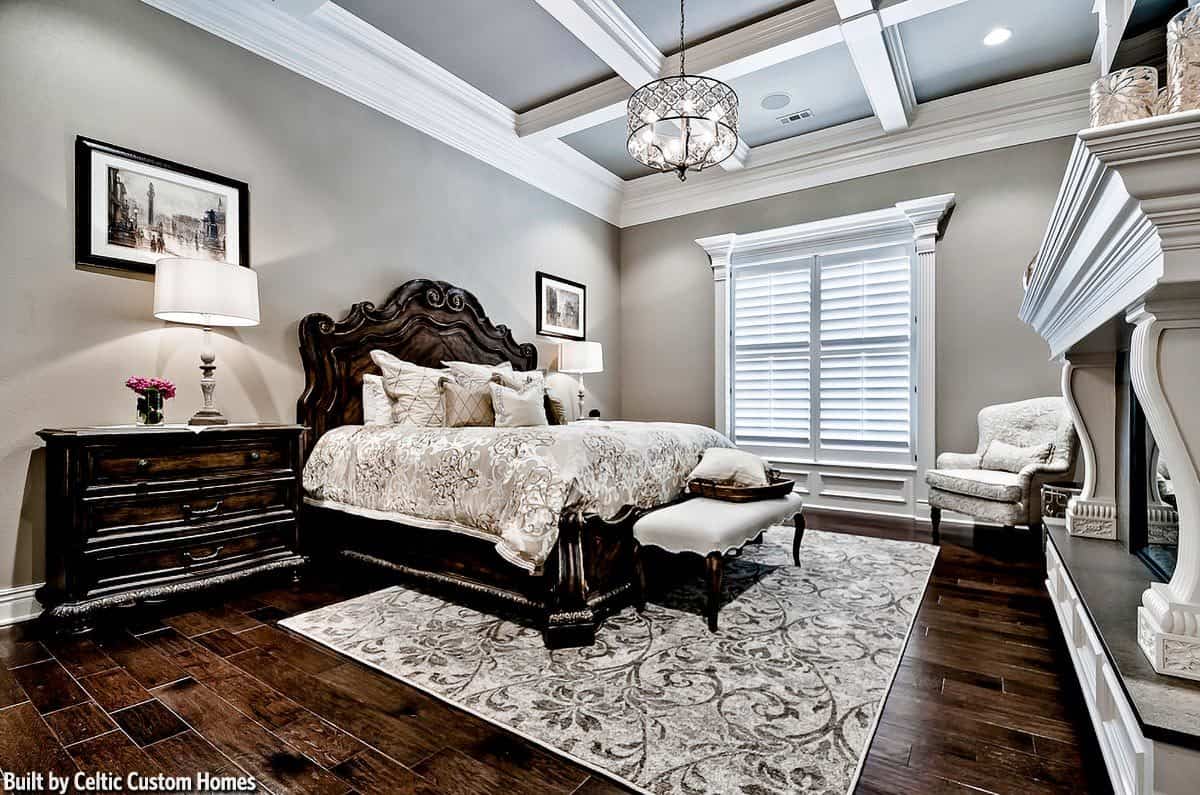 这间主卧的床对面有一座大壁炉，它的深色木质色调与床头抽屉相得益彰。这些与浅灰色的墙壁和格子天花板形成对比。