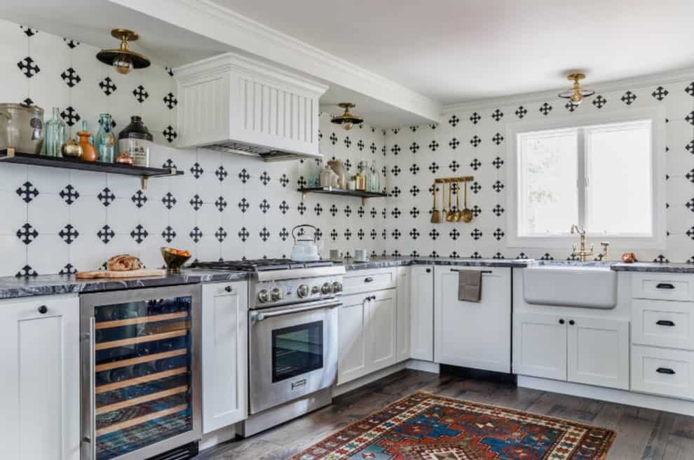 有图案的后挡板瓷砖在这个明亮的厨房中添加了鲜明的对比，白色橱柜和配有黄铜固定装置的农舍水槽。它有一扇玻璃窗和质朴的硬木地板，上面铺着印花地毯。