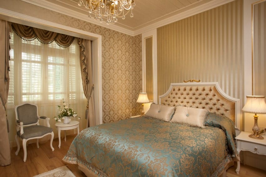 维多利亚风格的床的亮蓝色被子上有微妙的优雅的粉红色图案，与墙壁的粉红色图案壁纸以及美丽的簇绒床头板相辅相成，床头板被暖黄色的台灯照亮。