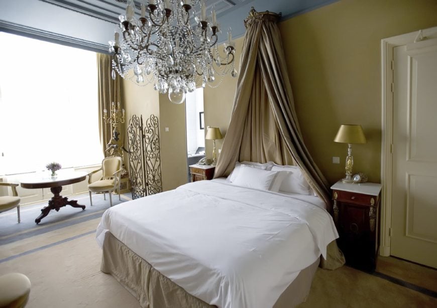 床头装饰的浅棕色窗帘与传统床的边裙相匹配，床的两侧是两个优雅的床头柜，白色的桌面上有金色的台灯。这些都与灰色的托盘天花板形成对比，天花板上有一盏明亮的水晶吊灯。