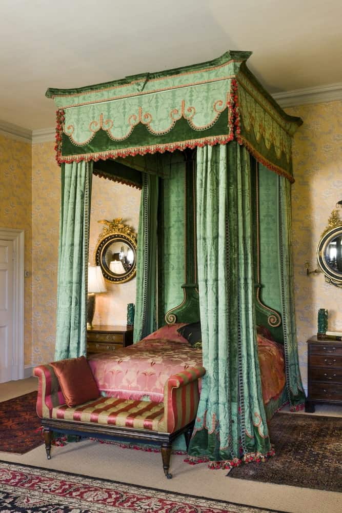这是一间色彩丰富的维多利亚风格卧室，有各种优雅的图案。这张四柱床上有红色图案的床单，与绿色图案的窗帘相映成辉。这些窗帘与有花卉图案的黄色花纹墙壁相映成趣。