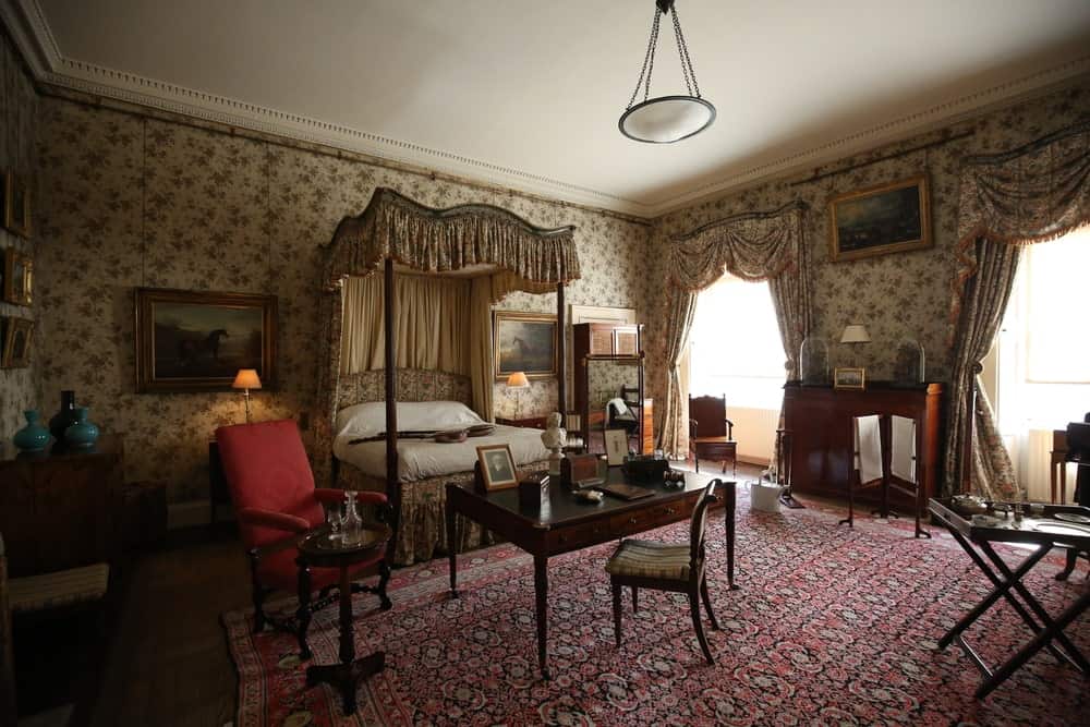 维多利亚风格的主卧室，有皇冠造型，室内壁纸，墙上有相框的照片，四柱床，大面积地毯上的座位区。
